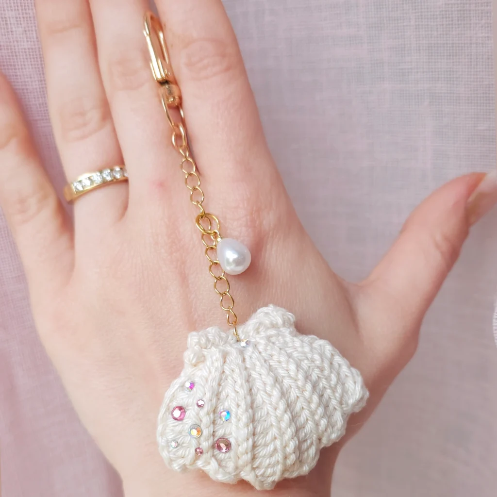 Joli bijou de sac composé d'un coquillage en crochet, orné de strass, sur une chaine dorée agrémentée d'une perle synthétique, fermoir mousqueton doré.